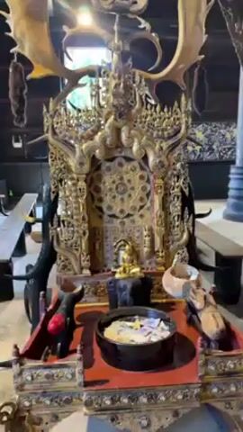 The camera pans around an interior shot of a dark room. The focus shifts from an ornately decorated altar in the center, adorned with a golden Buddha statue and surrounded by animal horns, to a large, light-colored wooden table. The table displays a variety of animal skins and skulls. In the background, an open door reveals a glimpse of greenery and blurred figures outside.
