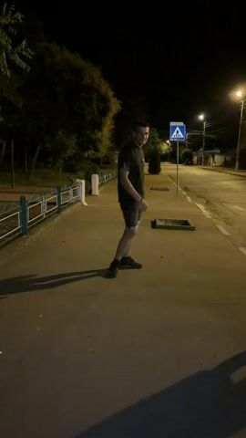 A young man wearing a dark t-shirt and shorts stands on a paved sidewalk at night. He is facing away from the camera and looking over his shoulder with a playful expression. The sidewalk is illuminated by streetlights, casting long shadows. To his left is a green fence and some trees. To his right is a street with a crosswalk sign. Another person's leg briefly enters the frame from the right side of the video, kicking towards the man. The man then gestures with his hand.
