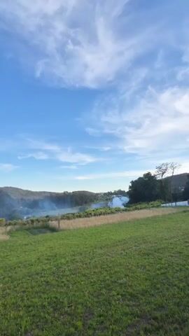 The video shows a field of green grass with a line of tall brown grass and a fence in the middle ground. Beyond that, there is a body of water covered in a layer of white smoke or fog. On either side of the water are hills or elevated land covered with green trees. The sky is bright blue with streaks of white clouds. In the center of the frame, fireworks are exploding in the sky, leaving a trail of white particles. The sound of fireworks can be heard.
