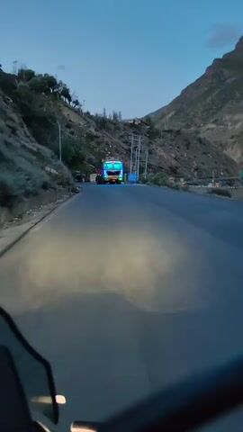 The video captures a journey on a mountain road at dusk. A large truck, illuminated by colorful lights, is prominently featured. The camera appears to be mounted on a vehicle, possibly a motorcycle, as evidenced by the reflection of headlights on the road. The surrounding environment is mountainous, with steep, rocky slopes and sparse vegetation. Several other vehicles, including what appears to be a white van, are visible in the distance. The sky is a deep blue, indicating the transition from day to night. The road is paved and appears to be in good condition. The overall atmosphere is serene and remote.