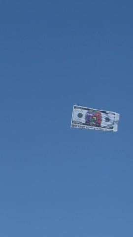 The video shows a large, flat object resembling a one hundred dollar bill floating in a clear, bright blue sky. The bill is rectangular and appears to be made of a light material, possibly paper or plastic. It's decorated with a colorful design in the center, partially obscuring the standard portrait, and includes the text "BEST GAME HERE". The bill is the main focus of the shot, contrasting starkly against the cloudless sky.