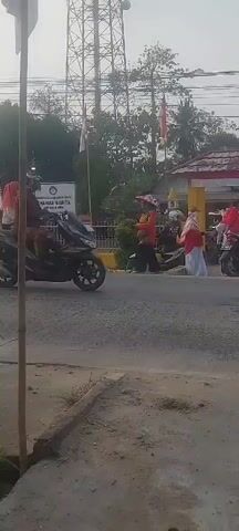 A group of people, mostly wearing red and white, are gathered outdoors. Some are on motorcycles, and others are walking. There are buildings in the background decorated with red and white banners, possibly indicating a celebration or event. One person is riding a black motorcycle, wearing a blue cap, white t-shirt with red sleeves, and black pants. Another motorcycle is carrying four people: a driver, two children in the middle, and a woman in the back wearing a white hijab and white shirt. The scene appears to be taking place during the daytime, with clear skies.
