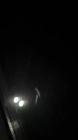 The video is shot from inside a dark vehicle looking out through a dirty windshield. Two bright, square headlights of an approaching vehicle are visible in the distance. The camera shifts slightly, and the headlights appear to grow dimmer before disappearing again. The background is mostly black with some light reflecting off the windshield.