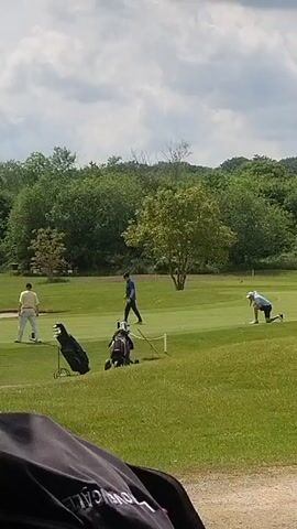 The video shows a golf course on a sunny day with some clouds. Three men are visible on the green, all engaged in playing golf. One man in a yellow shirt is standing next to a golf cart, another man in a blue shirt is walking towards the hole, and the third man in a white shirt is kneeling down. There are two golf bags and two golf carts on the green. The background features lush green trees and rolling hills. The video appears to be taken from inside a golf cart, as parts of the cart's interior are visible in the foreground.