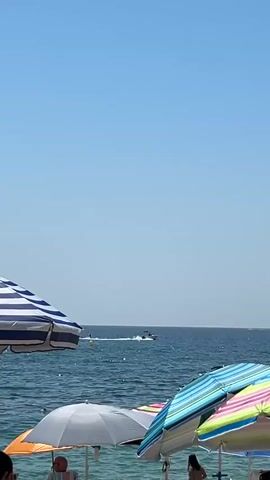 The video shows a beach scene with a clear blue sky. In the foreground, several beach umbrellas are visible, some with colorful striped patterns and others plain. In the middle ground, the calm blue ocean stretches out, with a small motorboat creating a wake as it moves across the water. A few yellow buoys are also visible in the water. In the background, the horizon is a faint line where the sea meets the sky. The overall atmosphere is one of a sunny day at the beach.