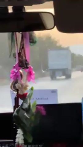The video, shot from inside a car, shows a view of a highway through the windshield. A large white truck is visible ahead, moving in the same direction. Several other cars are also present on the road, further in the distance. The foreground of the video features decorations hanging from the rearview mirror, including artificial flowers, ribbons, and woven green leaves. The car's dashboard is visible below, with a screen displaying a mirror image. The weather appears overcast, and the sky is a muted color.
