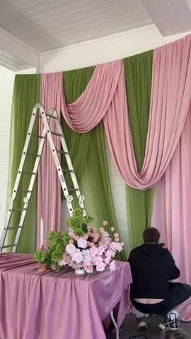 A medium shot shows a man in a black hooded sweatshirt kneeling and working on the bottom of a pink curtain panel in a room with white walls. To the left of the man is a silver metal stepladder, and a table covered in pink cloth. On the table is a floral arrangement consisting of green and pink flowers in a white pot, as well as a pink candlestick with a single white candle. Behind the man and to the left, the wall is adorned with alternating panels of green and pink fabric. The fabric is draped in a decorative swag pattern at the top.