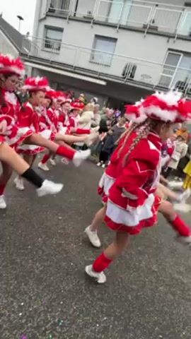 The video shows a group of young girls in red and white carnival costumes dancing in the street during a parade. They are wearing red jackets, short white skirts with red trim, red and white headwear, and white sneakers. They perform synchronized dance moves, including high kicks. Spectators line the street, watching and filming the performance. The buildings behind them appear to be European in style, with light-colored facades and multiple windows. The street is paved and there appears to be some litter. The sky is overcast.
