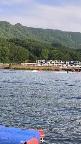 The video shows a wide shot of a body of water with a mountainous, tree-covered landscape in the background. A resort-like building with multiple floors and white balconies is visible along the shoreline. Several boats are in the water. In the foreground, a red and white inflatable tube is being pulled by a boat, with a person riding on it. Another boat is moving in the distance. The water is choppy, creating ripples on the surface. The sky is partly cloudy. The lower left corner of the frame shows a portion of a blue and orange floating platform.