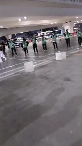 The video depicts a group of people, likely security personnel or event staff, forming a human chain under a bridge or overpass at night. They are wearing green vests and holding hands, presumably to control or direct the flow of pedestrian traffic. A group of people wearing traditional Islamic clothing can be seen walking past them. There are also concrete barriers visible in the foreground, likely used for crowd control. The overall environment appears to be an outdoor public space, potentially near an event or gathering, illuminated by streetlights.