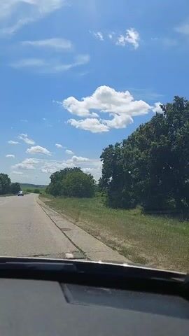 The video shows a view from inside a vehicle driving down a paved road on a sunny day. The road is bordered by green grass and trees on the right side, and trees and fields on the left side. The sky is blue with scattered white clouds. A car is visible in the distance on the left side of the road.