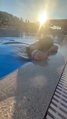 A person is floating in a swimming pool at sunset, with their head resting on the edge of the pool. The sun is setting in the background, casting a warm glow on the scene. The water is blue, and the pool deck is made of light-colored tiles. There are some trees and buildings in the distance. The person is wearing a dark-colored swimsuit.