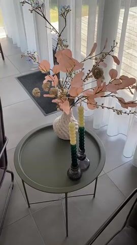 The video shows a close-up view of a decorative arrangement on a round, olive-green side table. The table is placed indoors, likely in a living area, with light filtering through sheer white curtains in the background. The scene is bright, suggesting it's daytime.

The main focus is a white, textured vase filled with a floral arrangement. The arrangement features branches with delicate light blue flowers and numerous large, soft pink leaves, possibly eucalyptus. Scattered throughout are small, pearl-like beads and some spiky, dried seed pods. Two tall, segmented candles in shades of light yellow and dark green are placed in dark, ornate candle holders on the table, to the right of the vase. The candles appear to be unlit.

The floor is tiled with large, light gray square tiles. To the left and right of the table, parts of dark-colored chairs are visible, hinting at a seating area. In the background, through the curtains, a glimpse of outdoor greenery and a door or window can be seen. A dark gray doormat with a subtle pattern is visible on the floor near the entrance in the background.

No people are visible in the video. The overall impression is one of a calm, stylish interior setting, possibly in a home. The lighting and decor suggest a peaceful and decorative ambiance. No visible text is present in the video.