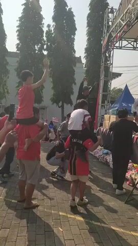 The video shows an outdoor event with a stage set up. Several people are gathered, with some on the stage and others in front of it. In the foreground, two individuals are giving piggyback rides to children. One man in a red shirt and grey shorts carries a child in a red shirt. Another person, wearing a red, black, and yellow outfit, carries a child in a white and red striped shirt. On the stage, a group of people appear to be performing or engaging in an activity, possibly singing or dancing, as some are holding microphones. In the background, trees and buildings are visible, suggesting an urban or semi-urban setting. The ground is paved with interlocking bricks. The overall atmosphere appears lively and festive.