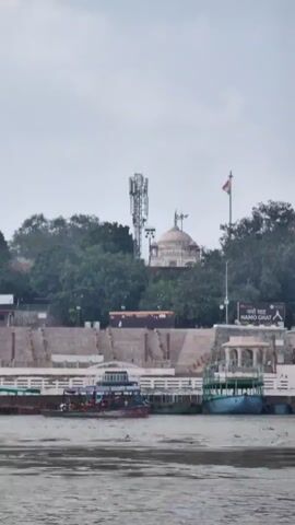A wide shot shows a body of water in the foreground and a cityscape in the background. Several boats are docked along the waterfront, and a large set of stairs leads up to a building with a domed roof. A cell tower is visible behind the building, and trees surround the area. The sky is overcast, and the overall mood is peaceful.
