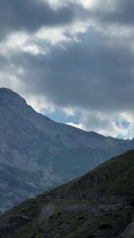 The video shows a view of a mountain range. The sky is cloudy, with some sunlight breaking through. The mountains are steep and rocky, with patches of green vegetation. A road or path can be seen winding its way up one of the slopes.