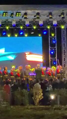 A choir stands on an outdoor stage at night, bathed in blue and yellow stage lights. They are led by a conductor. Behind them, a large screen displays nature scenes. A crowd watches from the front of the stage. A white van is parked on the street next to the venue.