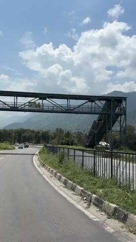 A vertical video shows a road with a pedestrian overpass and mountains in the background. The road curves to the left and is bordered by a fence and grass. A few vehicles are seen on the road. The sky is blue with white clouds.