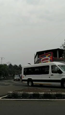 The video shows a street scene with a white van stopped at a corner. A smaller silver vehicle is driving on the street in the opposite direction. There is a billboard advertising Smirnoff behind the van, attached to a building. The sky is overcast. Palm trees and streetlights are visible along the road in the background.