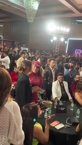 This video depicts a crowded event inside a large room with a high ceiling. Numerous people, mostly dressed in formal or semi-formal attire, are mingling and interacting. Some are seated at tables with water bottles and mobile phones, while others are standing and moving around. There are multiple photographers and videographers capturing the scene. 
Several women stand out with their unique outfits and hairstyles. In the foreground, a woman with red and black plaid clothing exchanges a kiss with a woman in a black coat. The room features lighting rigs overhead and a backdrop with a red and blue logo. The atmosphere seems lively and celebratory.