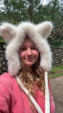 A young woman with blonde, wavy hair smiles at the camera while wearing a fluffy white hat with animal ears. She is wearing a pink hooded sweatshirt. The background shows a path, a stone wall, trees, and some red flowers. The video appears to be a selfie taken outdoors.