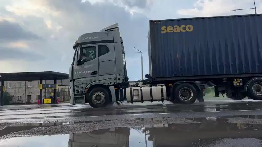 The video shows a large blue shipping container being transported by a semi-truck. The truck is driving on a wet road, with puddles reflecting the cloudy sky and surrounding buildings. The container has the word "seaco" written on its side. The truck appears to be in motion, indicated by the spray of water from its tires. In the background, there is a gas station with a prominent yellow logo and Cyrillic lettering, as well as other industrial-looking buildings. The overall scene suggests a logistics or transportation operation in an industrial or commercial area, possibly during or after rain.