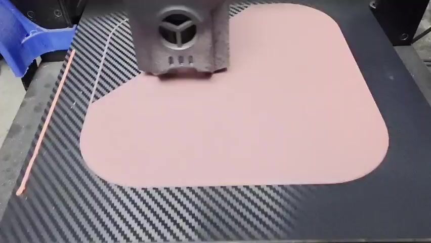 The video shows a close-up, high-angle view of a 3D printer's print head in action. The print head, a metallic gray device with visible vents and internal components, is positioned above a light pink, irregularly shaped object that is being printed on a black build plate. The build plate has a carbon fiber pattern. The print head is moving from left to right, and then appears to be tracing a curved line on the object. The object itself is partially printed, with the print head depositing material as it moves. In the background, a blue plastic component is visible, likely part of the 3D printer's structure. The overall environment appears to be a workshop or a makerspace.