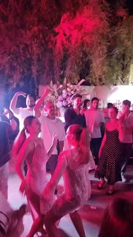 A group of people are dancing at an outdoor party. There are trees and foliage in the background. The lighting is red and purple, with a bright white light on the left side of the frame. Some of the people are wearing white shirts, while others are wearing more formal attire. The music is upbeat, and the people appear to be enjoying themselves.