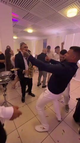 The video shows a lively wedding reception scene. The room has light-colored tile flooring and a white ceiling with recessed lighting, also confetti is scattered around. A musician in a black suit plays a saxophone. Several men, mostly in white pants and dark shirts, are dancing energetically around him. Some other people, including women, are visible in the background, some are dancing and others are watching. A set of drums is partially visible on the left side of the frame. The lighting in the room alternates between white, green, blue, and purple.