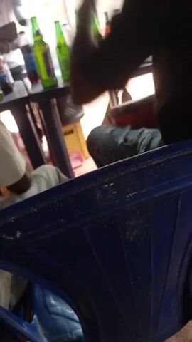 The video shows an outdoor gathering. Several people are seated around a black table with beer bottles and a soda bottle. The table is on a concrete surface, and the scene is viewed from behind a blue plastic chair. One person is wearing jeans and a black shirt, and another is wearing a white shirt. There is a yellow container and a pink object under the table. A person walks by in the background.