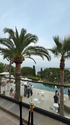 The video opens with a view of a resort or private residence with a swimming pool and palm trees. People are gathered by the pool, some standing and talking, others sitting at tables. The background features a cityscape with buildings and a body of water, likely the sea, with hills or mountains in the distance. The sky is overcast. A hand with a ring enters the frame and points towards the view. The camera then pans to show an arched doorway and large potted plants.