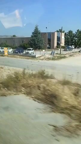 The video shows a view from a moving vehicle, likely a bus or car, looking out of a window. The foreground consists of dry grass and dirt, suggesting a somewhat arid environment. A paved road runs horizontally across the middle of the frame. On the other side of the road, there is a line of parked vehicles, including several white vans and some smaller cars. Further back, a long, low-rise building with a dark facade is visible. A tall green conifer tree stands in front of the building. To the right of the tree, a tall white sign with blue lettering is mounted on a pole. In the distance, a row of flagpoles with various flags is visible, and beyond that, rolling hills under a clear blue sky. The video captures a snapshot of an industrial or commercial area with some landscaping and infrastructure.