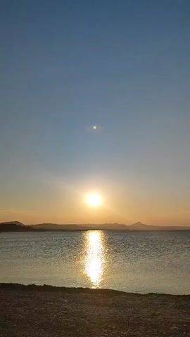 The video shows a sunset over a body of water. The sun is low in the sky, casting a bright reflection on the water. There are mountains visible in the distance. The sky is clear and blue. The foreground shows a beach with small pebbles. The video is static with no movement.