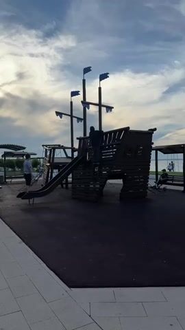The video captures an outdoor playground scene, likely in the late afternoon or early evening, given the soft, diffused light and partly cloudy sky. The main feature is a large, dark wooden pirate ship-themed playground structure with multiple masts adorned with blue flags. A black slide extends from the ship's platform to the ground. The play area is covered with a dark, rubberized safety surface, bordered by a white tiled pathway on the left.

Several people are present. One child, wearing a light-colored shirt, is seen climbing up the steps to the slide platform. Another child, in a dark shirt, is already on the platform of the pirate ship structure. An adult male in a light shirt stands near the base of the slide. In the background, two more individuals are visible, one seated on a bench under a covered structure. Beyond the playground, a large body of water, possibly the sea or a sizable lake, can be seen, indicating a coastal or lakeside setting. The overall atmosphere appears peaceful, with children engaged in play in a well-maintained recreational area. There is no visible text in the scene.