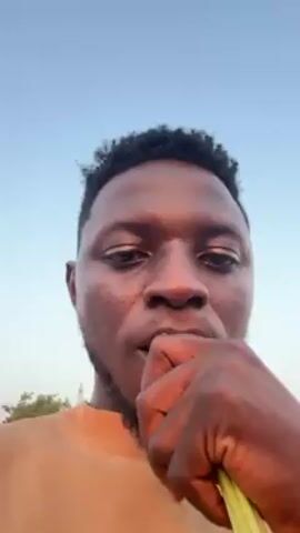 The video features a close-up shot of a man with dark skin and short, curly hair, wearing an orange shirt. He is chewing on a carrot with green stems still attached, looking directly at the camera.  The background appears to be an outdoor setting with a clear sky and trees visible. The camera angle is slightly upward, focusing primarily on the man's face.