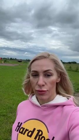 A blonde woman with makeup is filmed from the chest up in an outdoor setting. She is wearing a pink hooded sweatshirt with a "Hard Rock Cafe" logo. The background consists of a grassy field with some distant structures, including what appears to be a playground, and a cloudy sky. The woman's facial expression is neutral. The video is brief and consists of a series of short clips.