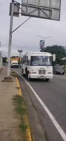 The video shows a white bus with "ENCANA" written on the front. A person is standing in the open doorway of the bus. The bus is on a road next to a sidewalk. There are other vehicles on the road, including a white truck and a green car. The sky is overcast. The video is shaky and appears to be recorded from a moving vehicle.