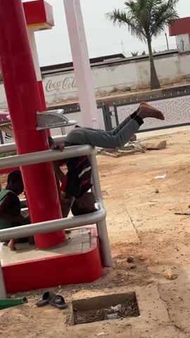 The video shows a young boy in a red and black shirt and grey pants hanging upside down from a metal railing. His head is facing the ground, and his feet are in the air. He appears to be supported by his arms, which are resting on the railing. Another child is sitting on the ground to the left of the boy. The background shows a red and white structure with the Coca-Cola logo visible, a red car, a palm tree, and a fence. The ground is covered with dirt and some debris. The overall scene appears to be outdoors, possibly at a public space like a park or a gas station.