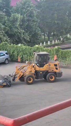 This media file shows a street sweeper machine in action on a paved surface. The machine is yellow and grey, with a large rotating brush at the front that is kicking up dust. Two cars are parked in the background, and a fence with decorative pillars runs along the edge of the paved area. Behind the fence, there are trees and foliage, suggesting an outdoor setting. A portion of a metal structure, possibly part of a crane or loading equipment, is visible in the foreground, with red safety railings. The lighting suggests it is daytime, possibly late afternoon or early evening. Visible text on the street sweeper includes "SLC" and the number "942." The metal structure in the foreground has blue text that reads "Rakoraf" and "PFEUFFER," and a yellow label with some text. The scene appears to be a municipal or commercial area, potentially a parking lot or a service area. No people are visible in the frame.