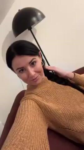 A woman with dark hair braided to the side is seated on a maroon colored couch. She is wearing a gold-colored turtleneck sweater. Behind her to the left is a black arc lamp with a dome shade. The woman looks directly at the camera and smiles.