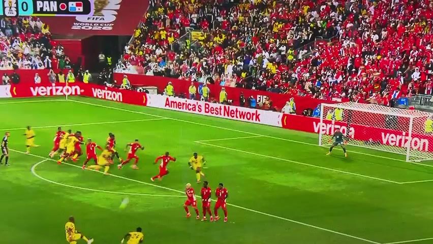 The video shows a soccer game. The field is green grass. There are two teams of players, one in red uniforms and the other in yellow uniforms. The players are running and kicking a soccer ball. The crowd in the stands is wearing red and yellow. There are advertisements around the field. The scoreboard shows 0 for both teams and "PAN" at the top left. The video is likely from a stadium during a professional soccer match.