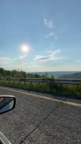 The video captures a view from a moving vehicle, looking out the passenger side window. The road is asphalt with a yellow line marking the lane. A metal guardrail runs along the side of the road, with lush green grass and trees behind it. In the distance, there are rolling hills and a bright sun shining in a clear blue sky with scattered clouds. The side mirror of the car is visible in the foreground, reflecting the sky and a hint of what appears to be a person's legs with pink shoes. The overall impression is a scenic drive on a sunny day.