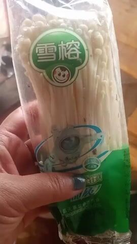 A person's hand holds a clear plastic package of enoki mushrooms. The package has green and white labeling with Chinese characters and a cartoon image of a smiling face. The mushrooms are white and long and thin. The background appears to be a wooden surface and possibly other blurred objects. The video is very short, showing the package from a slightly different angle each second.
