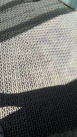 The video shows a close-up view of a textured metal surface, possibly a grate or mat, with a repetitive pattern. Sunlight casts shadows across the surface, creating a strong contrast between light and dark areas. The metal appears weathered and slightly dirty, with some visible debris or dirt caught within the openings of the pattern. In some sections, there are small circular metal elements that may be fasteners or part of the design. The overall impression is of an outdoor or industrial setting, with a focus on the textural details of the metal surface.