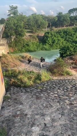 The video begins with a wide shot of two people standing on a small shore next to a river. The camera then focuses on one of them, who starts running up a stone-paved slope. The person is wearing a black sweatshirt with a graphic on the front, light-colored cargo pants, and sandals. They hold something in their hand as they run towards the camera. The background includes a green river, trees, and grassy areas. The slope is made of cobblestones.