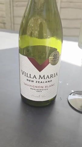 The video shows a bottle of Villa Maria New Zealand Sauvignon Blanc Marlborough 2023 wine on a dark surface, accompanied by a glass filled with the same white wine. The bottle is green with a white label and a gold seal indicating a 90-point rating. The wine glass has a silver stem. The background includes a blurred view of an outdoor setting. The camera shifts slightly, offering a full view of the bottle and glass. The background includes a wall. A voice, potentially of a child, speaks at one point.