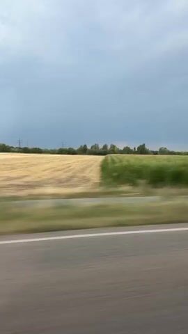 The video is a first-person perspective shot from a moving vehicle, likely a car, as indicated by the blurred foreground of a paved road. The scene unfolds horizontally, with the landscape streaking by. Dominating the frame is a vast field of green and yellow, appearing to be crops or tall grass, blurred due to the motion. Behind the field, a line of trees forms a distant horizon, interspersed with tall, slender structures that resemble electricity pylons. The sky above is overcast with a mix of grey and lighter blue clouds, suggesting a partly cloudy day. The overall impression is one of travel through a rural or agricultural area.