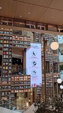 The video shows a large, multi-story bookstore with towering shelves filled with books. The architecture is modern, with wooden accents and open spaces. In the center of the frame, a large digital display shows various icons, including one of a person sitting, one of a person falling, and one of a person looking up. There are also signs with Korean text. People are seen browsing books, walking, and standing in groups. The overall atmosphere appears to be calm and inviting, with soft lighting.