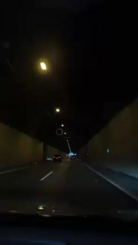 The video shows the perspective from inside a car driving through a tunnel. The tunnel has concrete walls and a dark ceiling with evenly spaced lights. The road is divided by a solid white line, and other cars are visible ahead in the same lane. The light at the tunnel's end appears white.