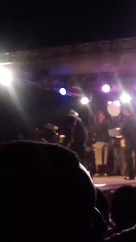 The video shows a performance stage lit with bright white and purple lights. The view is from the audience looking up at the stage. Several figures, presumably performers, stand on the stage, though their faces are not visible. Some of the audience members hold up phones, presumably to record the performance. There are stage lights and a truss system above the performers.